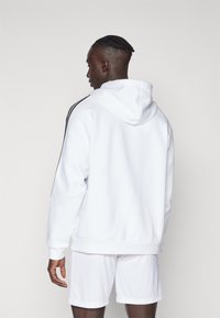 Weiße Kapuzenpullover mit lässiger Passform, ausgestattet mit schwarzen Seitenstreifen und einer Kängurutasche, kombiniert mit weißen Shorts. Glatte Textur.