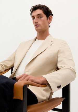 BlazerBeige de tela ligera, combinado con una camisa blanca. Sentado en una silla con reposabrazos marrones, con un fondo neutral.