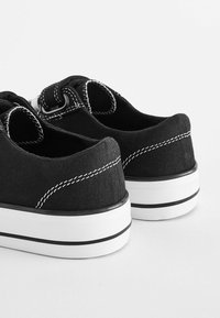Sorte kanvas sneakers med hvid syning og detaljer, rund tå, lav profil og gummisål med sorte og hvide vandrette striber.