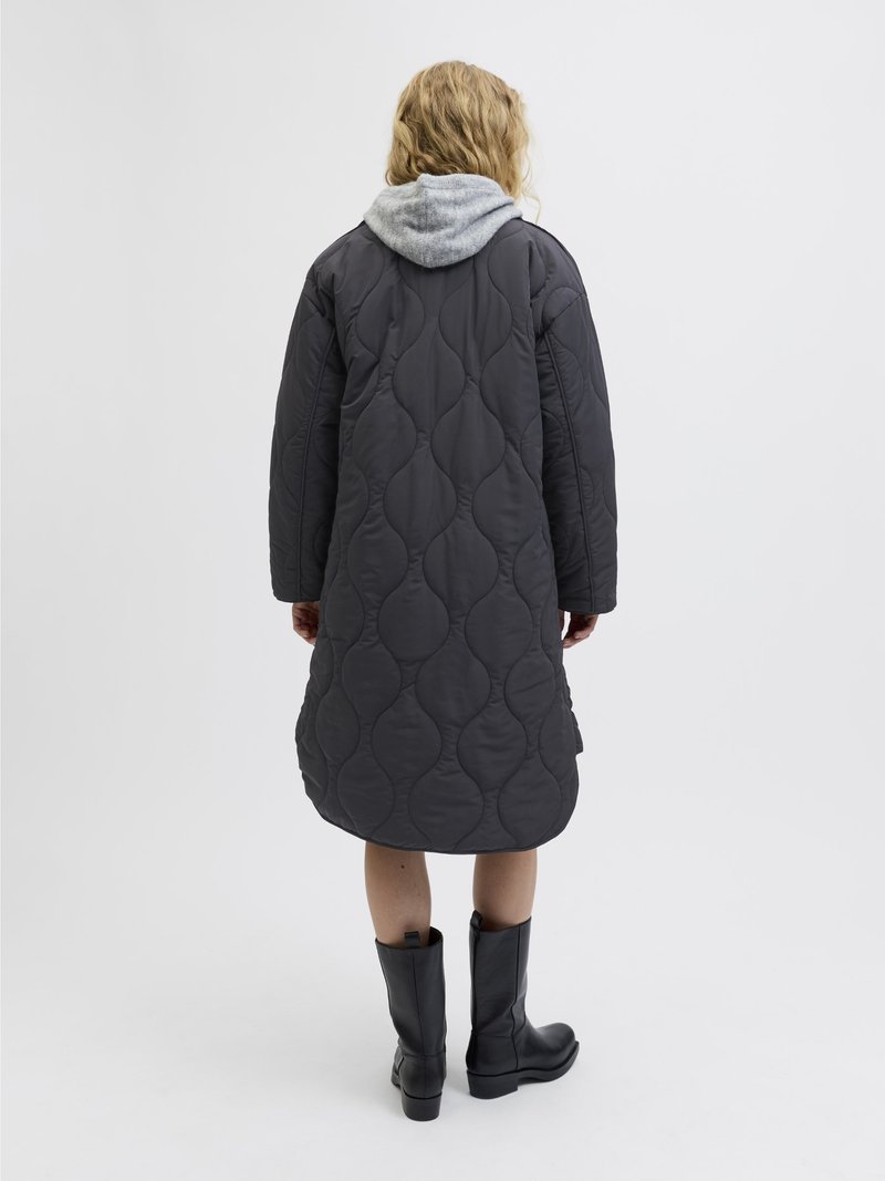 Langer, gesteppter Mantel in Dunkelgrau mit einem strukturierten Muster. Verfügt über eine graue Kapuze und einen abgerundeten Saum, kombiniert mit knielangen schwarzen Stiefeln.