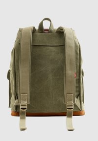 Πράσινο σακίδιο πλάτης από καμβά με καφέ υφασμάτινο κάτω μέρος, ρυθμιζόμενους ιμάντες και χειρολαβή στο πάνω μέρος. Διαθέτει μια επίπεδη τσέπη στην πλάτη.