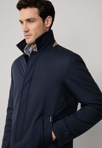 Navyblauwe jas met een hoge kraag, voorzien van een grijze binnenvoering, beige accenten op de kraag en een zijzak met een logo.