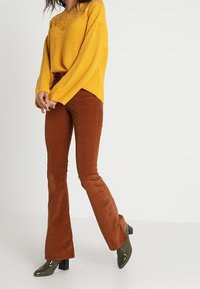 Musztardowy sweter z dzianiny z koronkowym dekoltem, zestawiony z brązowymi spodniami dzwonami. Modelka nosi zielone botki na obcasie, prezentując casualowy wygląd.