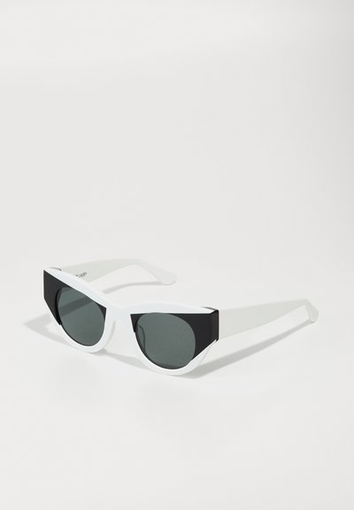 Lunettes de soleil en forme de chat avec une monture supérieure noire mate et une monture inférieure blanche brillante. Les verres sombres et la texture lisse ajoutent au design moderne.