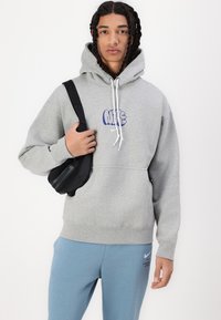 Šedá mikina s kapucí a modrým logem Nike, s tkaničkami na kapuci a klokaní kapsou. Kombinováno s modrými tepláky a černou taškou přes rameno.