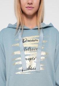 Hanorac de culoare albastru deschis, cu șnur, având un design mare din text metalic auriu și negru cu cuvintele "Visiți", "Credeți", "Inspirați", "Realizați."