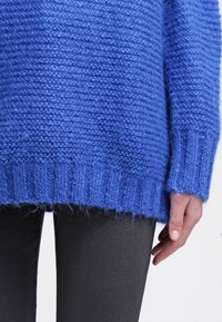 Gros plan sur une personne portant un pull en tricot bleu texturé et un pantalon noir, avec une main posée à côté de la hanche.