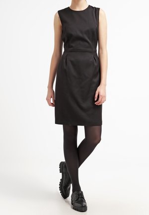 Mujer de pie con las piernas cruzadas, lleva un vestido negro sin mangas, medias opacas negras y zapatos negros gruesos con cordones.