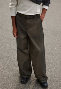 Pantalons à jambes larges en tissu olive foncé, avec une texture lisse, une taille ajustée et des détails de pli subtils. Associés à des chaussures noires.