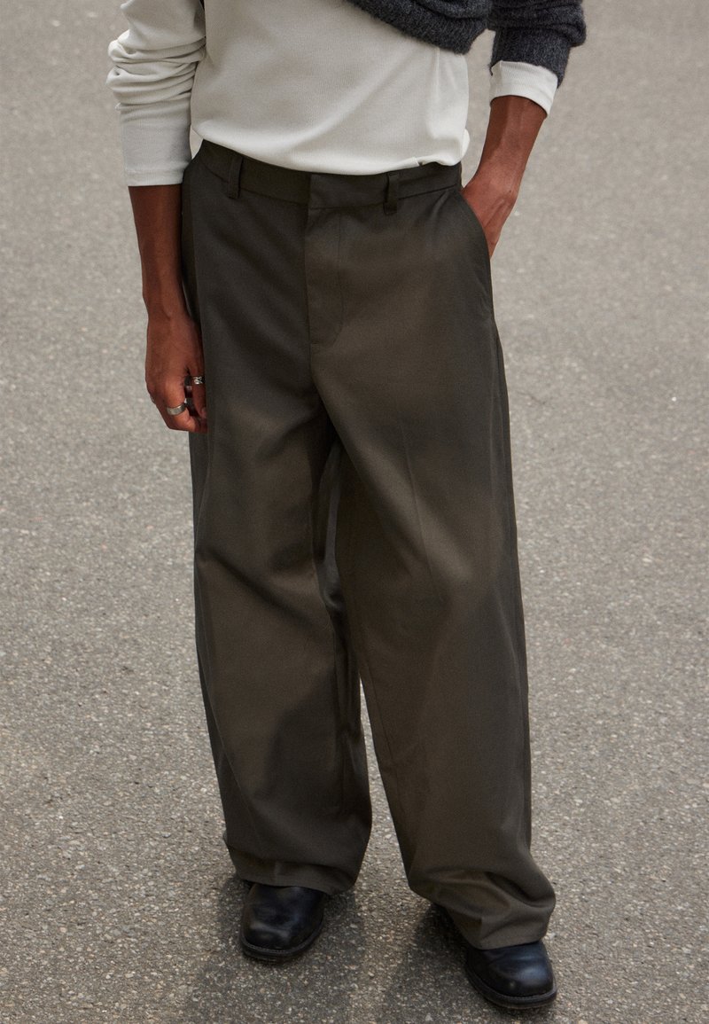 Pantalons à jambes larges en tissu olive foncé, avec une texture lisse, une taille ajustée et des détails de pli subtils. Associés à des chaussures noires.