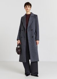 Cappotto doppio petto grigio in lana con rever a punte, bottoni neri e tasche laterali, indossato sopra una maglietta bordeaux e pantaloni neri larghi.