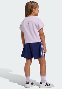 Svetlofialové tričko s krátkym rukávom, tmavomodré šortky s bielymi pruhmi, skombinované s bielymi teniskami a svetlofialovými ponožkami. Na tričku je logo Adidas.