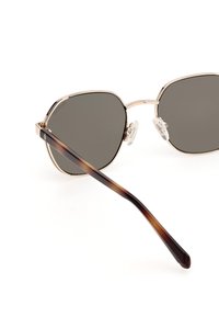 Gafas de sol con marco de metal bronce y lentes grises, que presentan una forma hexagonal única, una varilla de carey y almohadillas nasales transparentes.