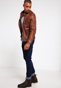 Chaqueta de cuero marrón con detalles de cremallera, combinada con una camisa gris, jeans oscuros y botas marrones. Textura suave y diseño ajustado.