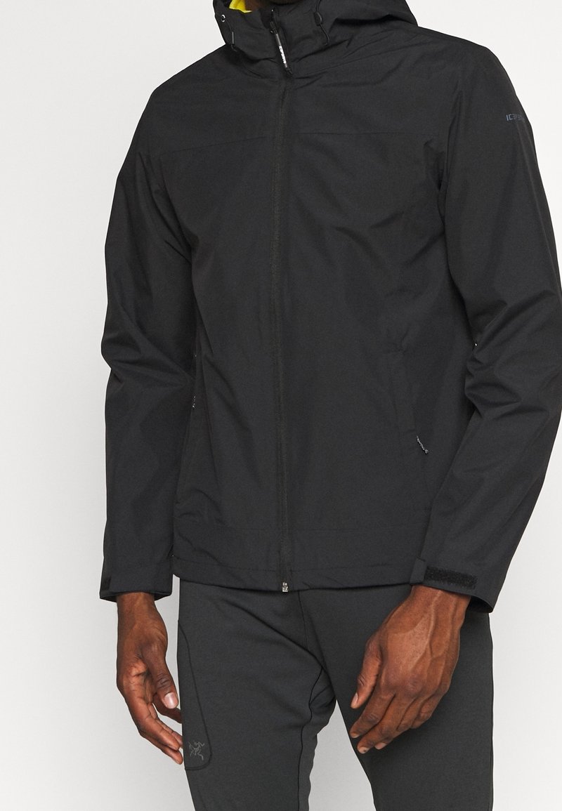 Homme portant une veste noire à capuche avec fermeture éclair et un pantalon noir, debout les bras détendus le long du corps.