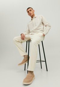 Striped, long-sleeve shirt in white and light brown; cream pants; tan slip-on shoes; seated on a black stool with a minimalist background.