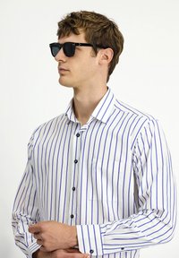 Camicia bianca con strisce verticali blu, maniche lunghe, colletto button-down, taschino frontale e bottoni neri. Occhiali da sole neri indossati.