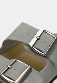 Γκρι σουέτ σανδάλια με δύο ρυθμιζόμενους μεταλλικούς σφιγκτήρες, ανοιχτές πλευρές και καφέ δερμάτινη σόλα. Μαλακή υφή με μινιμαλιστικό σχέδιο.