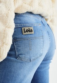 Jeans in denim blu con una texture sbiadita, caratterizzati da una toppa rettangolare in pelle grigia con il marchio "Lois" sul retro.
