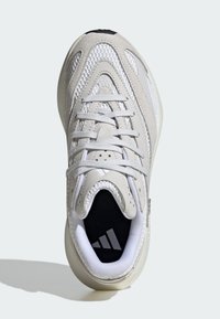 Zapatilla gris claro con parte superior de malla, acentos de ante texturizado, cordones blancos y un logo negro en una lengüeta acolchada. Cuenta con una suela suave en color crema.