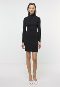 Svart langermet, tettsittende kjole med høy hals, laget av et glatt stoff, kombinert med hvite spissede hæler. Enkel og elegant design.