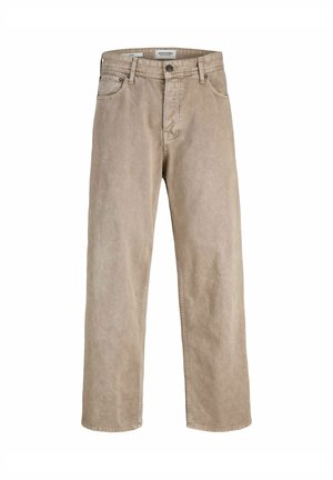 Personne portant un t-shirt noir avec le texte "Originals Studio", un pantalon beige à jambes larges et des baskets blanches, debout devant un fond uni.