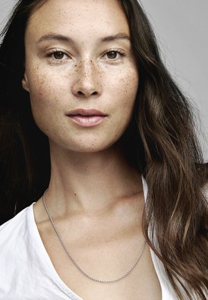 Collana di argento appoggiata su un collo nudo, abbinata a una camicia bianca. La pelle presenta sottili lentiggini e una texture liscia.