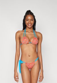 Il set bikini presenta un tessuto rosso brillante con motivi floreali multicolori, spalline blu regolabili e una parte inferiore con lacci per una vestibilità personalizzata.
