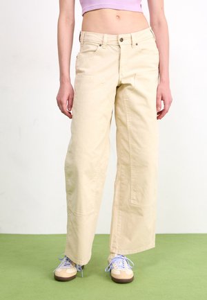Persona que lleva pantalones anchos beige, top corto morado y zapatillas crema con cordones azul claro, de pie sobre suelo verde.