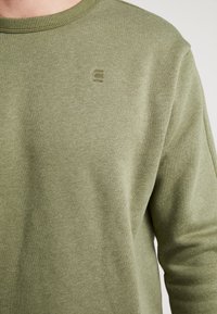 Jersey verde oliva hecho de una tela suave, con un cuello acanalado y un pequeño logo bordado en el pecho. Superficie simple y texturizada.