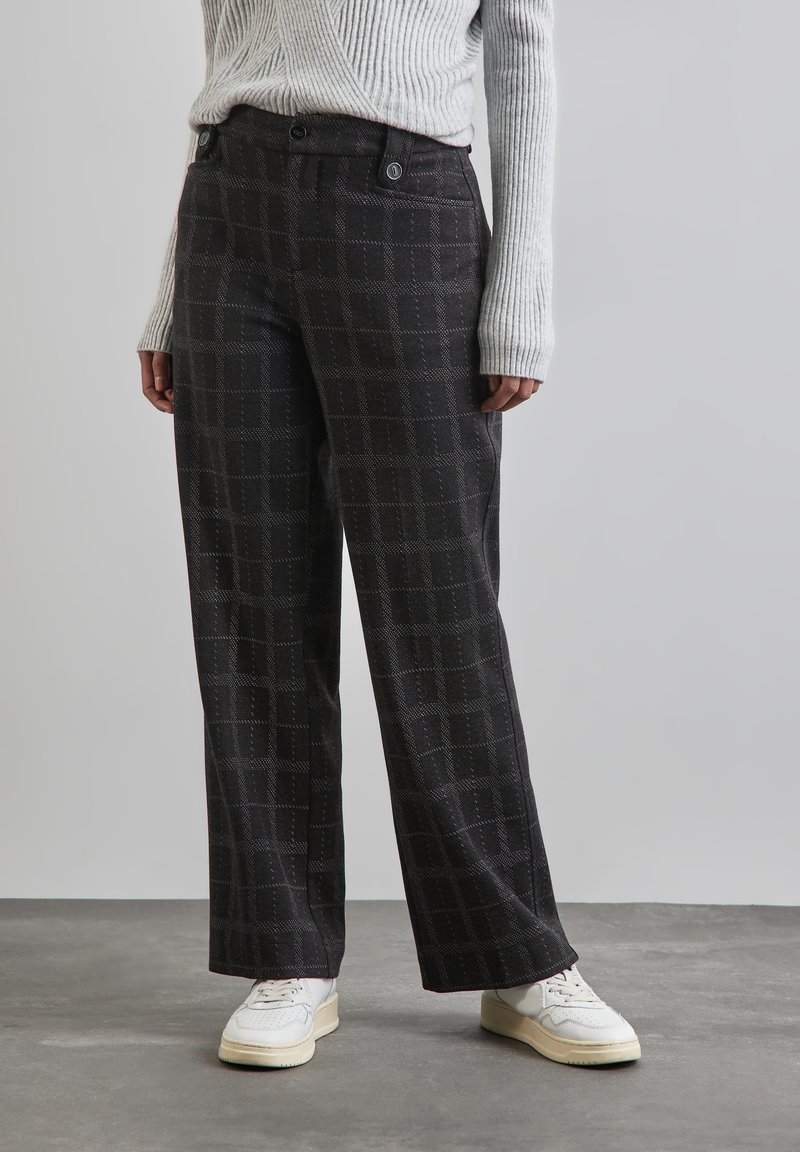Schwarze weit geschnittene Hose mit einem dezenten Karomuster. Der Stoff hat eine strukturierte Oberfläche und verfügt über zwei Knöpfe an der Vorderseite. Getragen mit weißen Sneakers.
