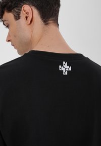 Black cotton T-shirt featuring a round neckline and a white geometric logo printed at the back near the shoulder. Soft texture.