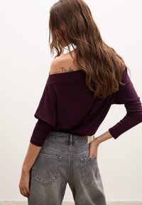 Top a maniche lunghe, scollato sulle spalle in un profondo viola abbinato a jeans grigi sbiaditi a vita alta. Presenta una vestibilità comoda e una texture morbida.