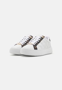 Baskets blanches avec une tige texturée, présentant un design matelassé, des accents latéraux noirs à motifs, et des semelles épaisses et rainurées. Lacets blancs.