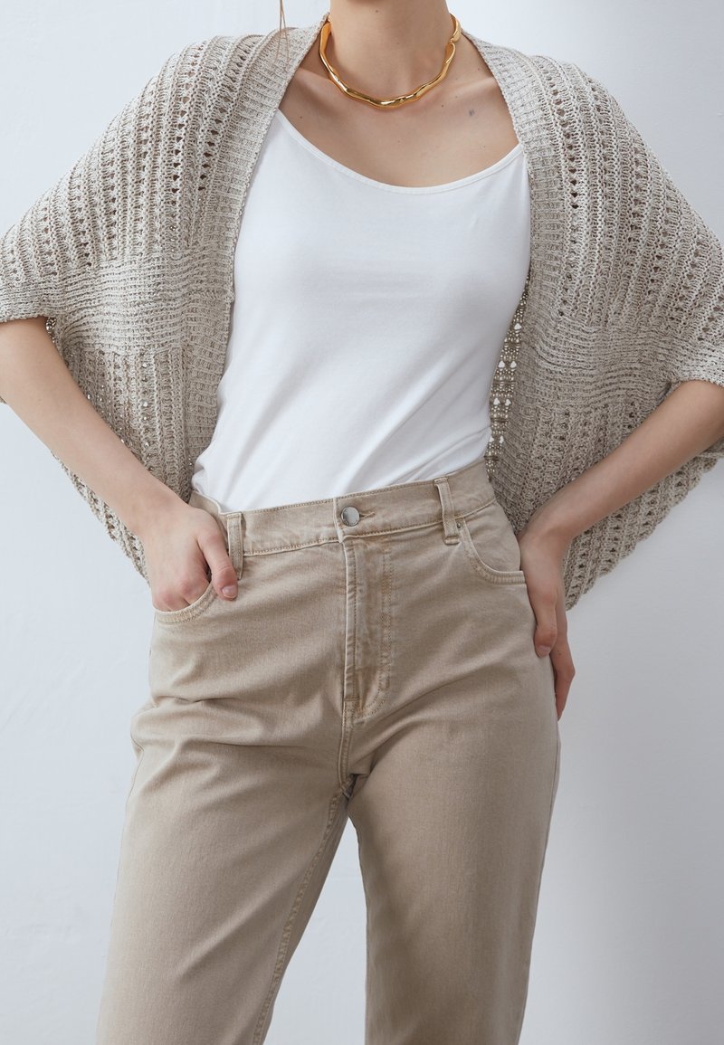 Cardigan en tricot beige clair à devant ouvert et manches larges, porté sur un haut blanc et assorti à un pantalon taille haute beige clair.