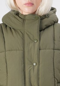 Dukati maslinasto zeleni puffer prsluk s visokim ovratnikom, kopčama na dugmad i glatkom teksturom. Sadrži elastične podešivače na kapuljači.