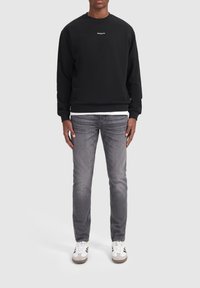 Zwarte sweatshirt met ronde hals en wit logo, gecombineerd met grijze jeans en witte sneakers met grijze accenten. Glad materiaal en casual pasvorm.