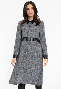 Schwarz-weißes Hahnentrittmuster-Kleid mit langen Ärmeln, einem Kragen und Akzenten aus Kunstleder an den Bündchen und der Taille. Knopfleiste vorne.