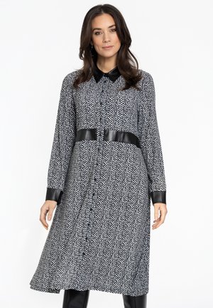Robe en pied-de-poule noir et blanc à manches longues, avec un col et des accents en simili cuir aux poignets et à la taille. Fermeture à boutons sur le devant.