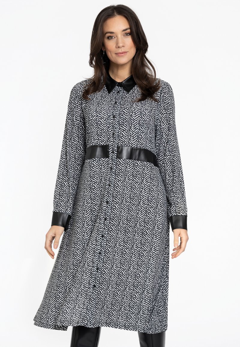 Schwarz-weißes Hahnentrittmuster-Kleid mit langen Ärmeln, einem Kragen und Akzenten aus Kunstleder an den Bündchen und der Taille. Knopfleiste vorne.