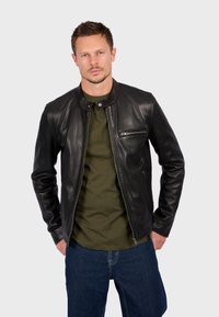 Chaqueta de cuero negra con cuello alto, cierre frontal de cremallera y un bolsillo en el pecho con cremallera. Lleva puesta una camisa de color verde oliva.