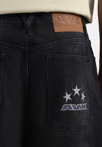Pantalones de mezclilla negros con un bolsillo trasero que presenta estrellas bordadas en blanco y el logo "AVX". Parche de cuero en la cintura con texto en relieve.