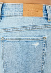 Ljust blå denimjeans med en läderetikett vid midjan, synlig sömnad och ett slitet patch på bakfickan.