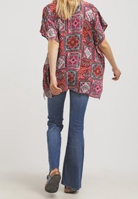 Haut de style kimono à motifs rouges et roses avec des manches courtes, associé à un jean bleu évasé et des sandales noires. Tissus texturés et motifs géométriques.