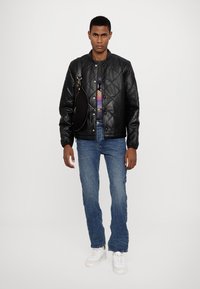 Veste bomber matelassée noire, chemise graphique multicolore, jean bleu, baskets blanches, sac à bandoulière noir rond. Tenue simple et décontractée.