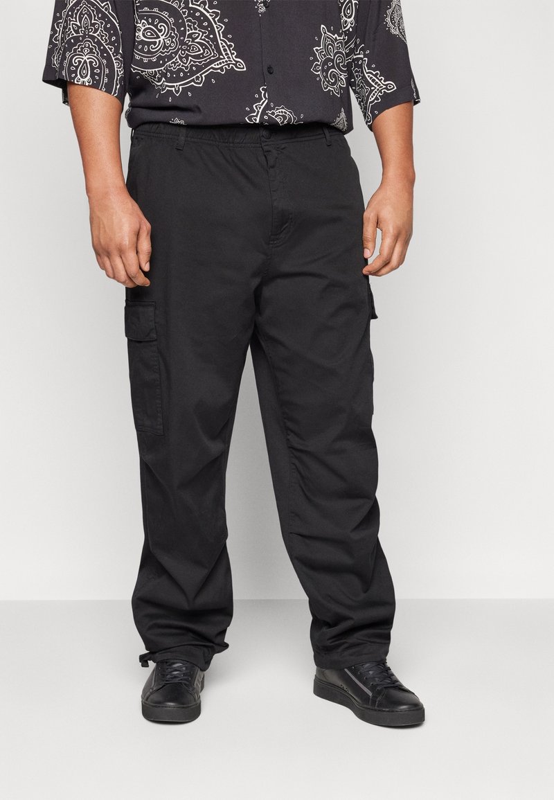 Mies, joka on pukeutunut mustiin cargo-housuihin, mustiin tennareihin ja mustaan paitaan, jossa on valkoisia paisley-kuvioita, seisoo yksinkertaista taustaa vasten.