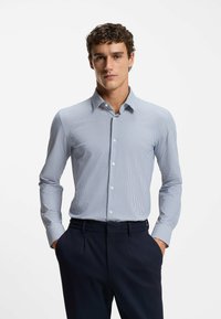 Lichtblauw-wit geruit overhemd met een button-down kraag en lange mouwen. Gecombineerd met een donkere broek, handen in de zakken.