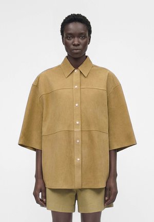 Άτομο με σκουρόχρωμο δέρμα φοράει υπερμεγέθη μπεζ πουκάμισο με φαρδιά μανίκια και ταιριαστά σορτς σε μονόχρωμο φωτεινό φόντο.