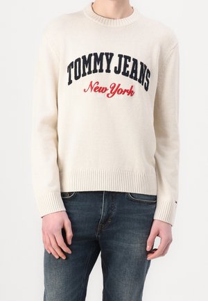 Henkilö, joka pukeutunut kermaanväriseen neulepuseroon, jossa lukee "TOMMY JEANS New York" rinnassa, ja tumansiniset farkut, vaaleaa yksinkertaista taustaa vasten.