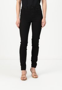 Black skinny jeans with a classic five-pocket design, crafted from denim, featuring belt loops and a zip fly, paired with beige strappy heels.